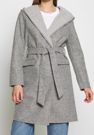 Frau, die einen grauen Wollmantel mit Gürtel und breiten Revers über einem weißen Oberteil und blauen Jeans trägt, vor einem schlichten Hintergrund stehend.