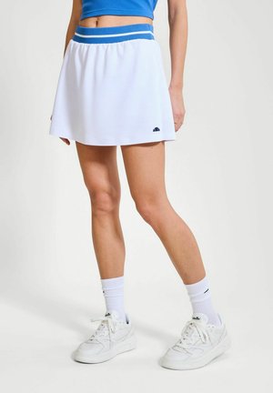 Frau trägt einen weißen Tennisrock mit blauem Bund, weiße Socken und weiße Turnschuhe, steht vor einem schlichten hellen Hintergrund.