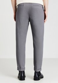 Pantaloni grigi su misura con una texture liscia, caratterizzati da orli arrotolati e tasche posteriori. Indossati con scarpe in pelle nere.