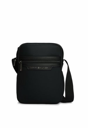 Kleine schwarze Tommy Hilfiger Umhängetasche mit vorderer Reißverschlusstasche und verstellbarem Schulterriemen auf weißem Hintergrund.