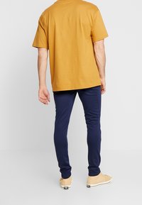 Bavlněné hořčicové tričko s krátkým rukávem v kombinaci s tmavě modrými slim-fit kalhotami a béžovými teniskami, zobrazené jako ležérní outfit zezadu.