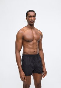 Άνδρας χωρίς πουκάμισο με μυώδες σώμα που φοράει μαύρα μποξεράκια, στέκεται μπροστά σε απλό ανοιχτό γκρι φόντο.