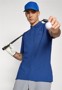 Kék férfi póló, rövid ujjú, textúrált anyagból, gombos záródással. Golf labdát és ütőt tart, kék sapkát és fehér kesztyűt visel.