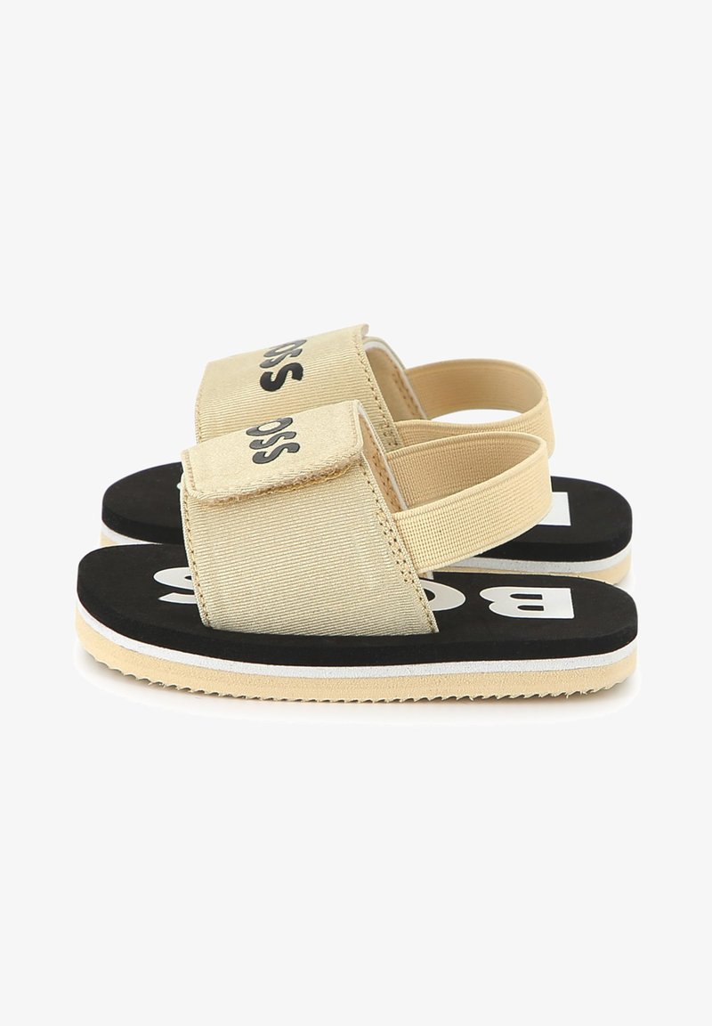 Beige fabric sandals with a velcro strap, black sole featuring white accents, and a textured footbed. Logo displayed in black on the strap.
