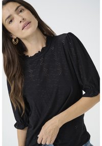 Blusa nera in tessuto texturizzato con scollatura arricciata e maniche a sbuffo. Presenta modelli intagliati al laser su tutta la superficie.