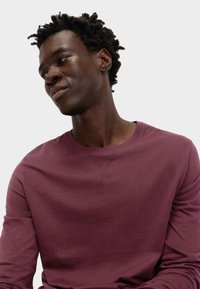 Camisa de manga larga burdeos en tejido suave con cuello redondo y detalle de costura visible. El modelo está ligeramente de lado.