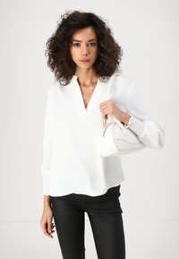 Valge popover-teks koos tekstureeritud kangaga ja V-kaelusega. Mustad slim-fit püksid. Modell hoiab käes valget kumerat käekotti.