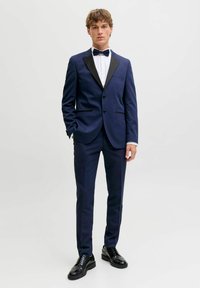 Giovane uomo in piedi che indossa uno smoking blu navy con rever neri, camicia bianca, papillon nero e scarpe eleganti nere su uno sfondo semplice.