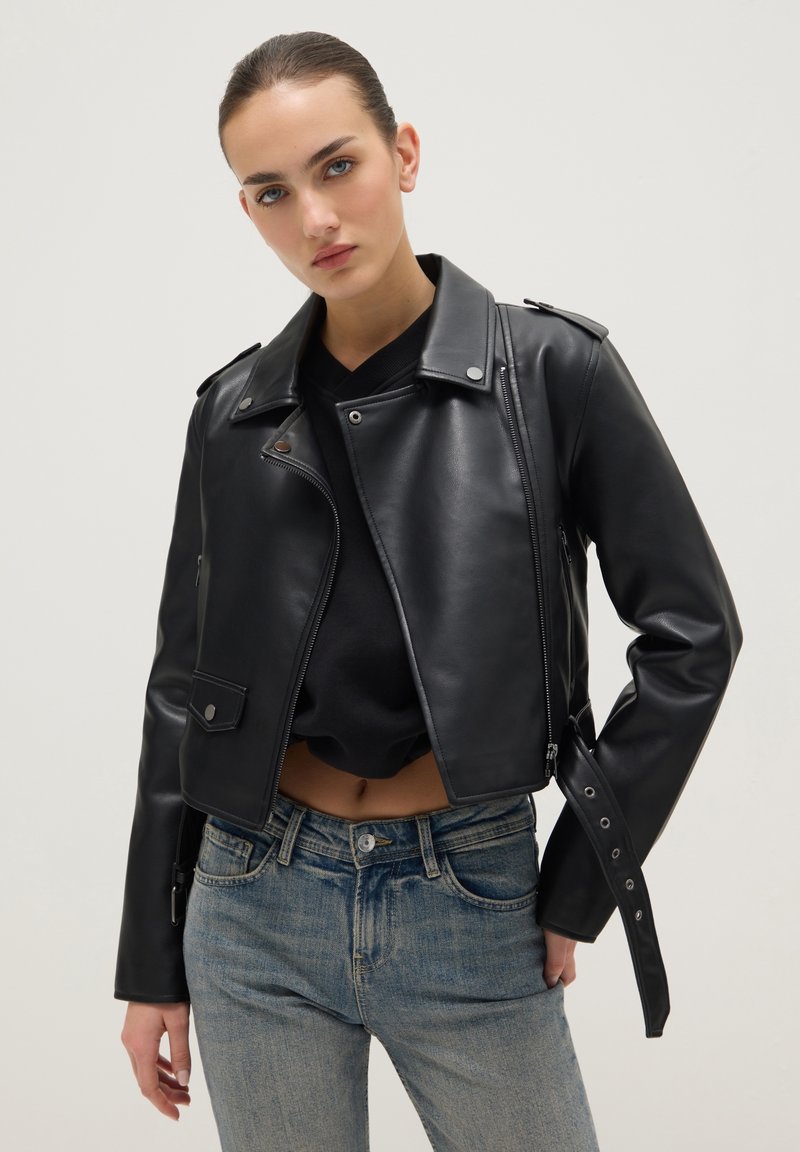 Zwarte cropped leren jas met epauletdetail, ritssluiting aan de voorkant en zakken met drukknoop, gedragen over een zwart shirt en blauwe spijkerbroek.