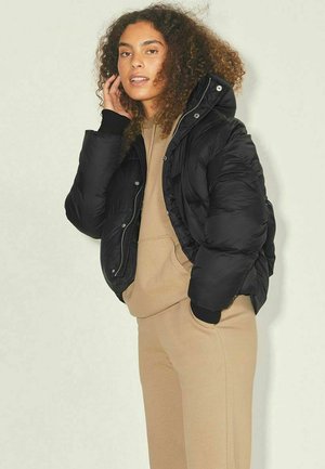 Black puffer jacket with a zippered front, high collar, and ribbed cuffs, worn over a beige hoodie, against a neutral background.