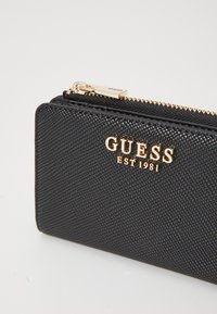 Portafoglio nero texturizzato con chiusura a zip, dotato del logo dorato "GUESS EST 1981" sulla parte anteriore. Bordo liscio e forma rettangolare compatta.