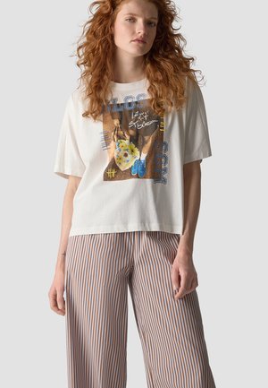 Frau mit lockigem roten Haar, die ein weißes grafisches T-Shirt und braun-weiß gestreifte weite Hosen vor einem schlichten Hintergrund trägt.