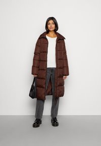 Brauner gepolsterter Mantel mit großem Kragen, langen Ärmeln und mittellangem Design, getragen über einem weißen Oberteil und kombiniert mit dunkelgrauen Jeans.