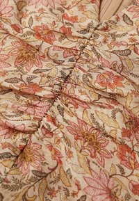 Détail de tissu floral avec un fond beige et des fleurs roses, jaunes et brunes. Texture froncée avec des accents de couture élégants.