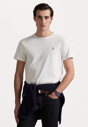Tricou alb din bumbac cu guler rotund, având un mic logo brodat. Asortat cu blugi închiși la culoare și un pulover bleumarin legat în jurul taliei.