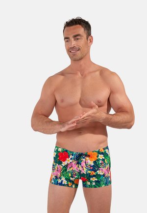 Hemdloser Mann mit kurzen lockigen Haaren, der bunte florale Badeshorts trägt, lächelt und reibt seine Hände gegeneinander vor einem einfarbigen Hintergrund.