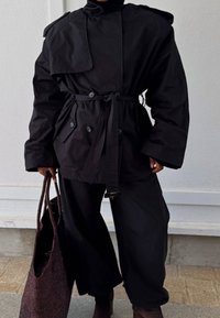 Czarna kurtka z zewnętrzną powłoką, z paskiem w talii, podwójnymi guzikami z przodu i dużymi klapami. Połączona z luźnymi czarnymi spodniami i ciemną tkanym torbą.