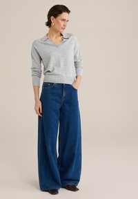 Siv niahtani pulover s ovratnikom in rebrastimi manšetami, v kombinaciji s širokimi temno modrimi jeans hlačami. Obleka ima sproščen kroj.