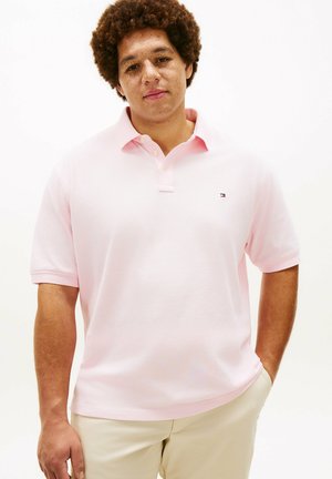 Ung mand med krøllet hår iført en lyserød polo-shirt og beige bukser, stående med den ene hånd i lommen på hvid baggrund.