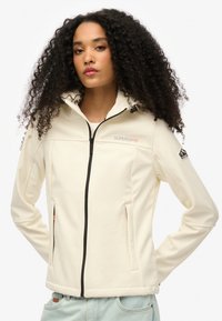Witte softshelljack met een hoge kraag, ritssluiting en twee zijzakken. Bevat het Superdry-logo en een bergpatch op de mouw.