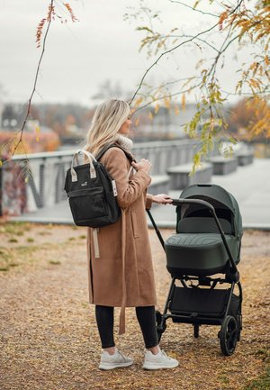 Woman in beige coat with black backpack pushing dark green baby stroller on autumn park path near metal railing bridge.