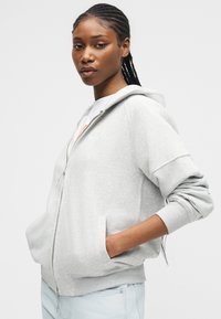 Mladá žena s pletenými vlasmi, oblečená v svetlosivej mikine so zipsom a svetlomodrých džínsoch, stojaca s jednou rukou v vrecku.