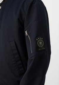 Dark blue jacket sleeve with a zippered pocket and a patch featuring four stars, an eagle emblem, and "Marc O'Polo" text.