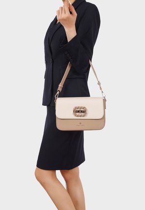 Donna in completo business blu navy che tiene una borsa a tracolla beige e crema con dettaglio a catena sulla chiusura.