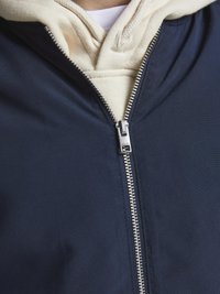 Námořnická zipová bunda přes krémovou mikinu s kapucí. Obsahuje stříbrný zip a hladkou, texturovanou tkaninu. Kapuce s viditelnou šňůrkou.