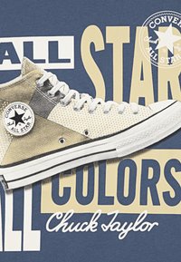 Beige visokotopni Converse Chuck Taylor superge z belimi akcenti, pikčasto teksturo in črnimi detajli na modri podlagi s besedilom.