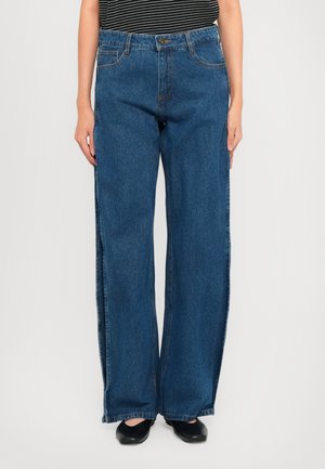 Person som har på seg løstsittende, blå denimjeans, svarte flate sko og en svart-hvit stripete ermeløs topp, står mot en hvit bakgrunn.