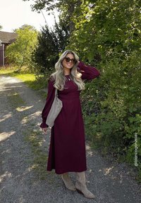 Robe bordeaux à manches longues avec une taille cintrée et une jupe fluide. Assortie à des bottines beiges à la cheville et un sac à épaule marron clair. Fond de feuillage vert.