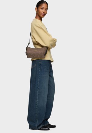 Femme aux cheveux tirés en arrière portant un pull beige et un jean bleu à jambes larges, portant un petit sac à bandoulière marron à motifs.