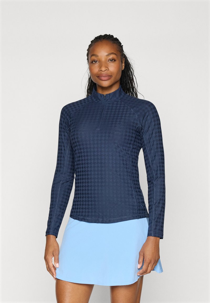 Marineblå langermet topp med høy krage og delvis glidelås, med et teksturert houndstooth-mønster. Kombinert med et lys blått skjørt.