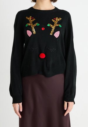 Pull noir avec des bois de renne en sequins dorés, des oreilles roses et un nez en pompon rouge, porté par une personne en jupe marron, fond blanc.