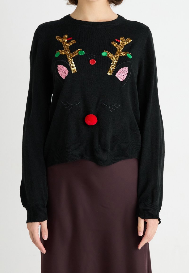 Pull noir avec des bois de renne en sequins dorés, des oreilles roses et un nez en pompon rouge, porté par une personne en jupe marron, fond blanc.