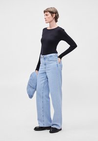 Ανοιχτό μπλε φαρδύ τζιν με ψηλή μέση, συνδυασμένο με μια μαύρη ριγέ μπλούζα μακρυμάνικη. Κρατάει ένα τζιν μπουφάν. Μαύρα παπούτσια.