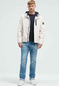 Giacca impermeabile bianca con dettagli neri e cappuccio, indossata sopra una maglietta scura, abbinata a jeans azzurri chiari e sneakers bianche.