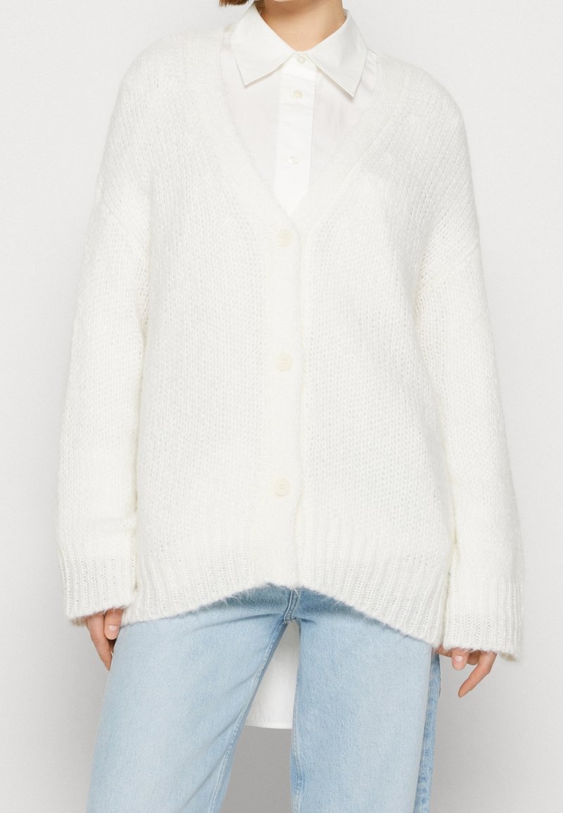 Femme portant un cardigan blanc tricoté surdimensionné avec des boutons, par-dessus une chemise blanche à col, et un jean bleu clair, debout devant un fond uni.
