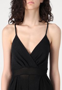 Crna haljina bez rukava s dubokim V-izrezom i dizajnom omotanja. Izrađena od teksturirane, reflektirajuće tkanine s uskim strukom i lepršavom suknjom.