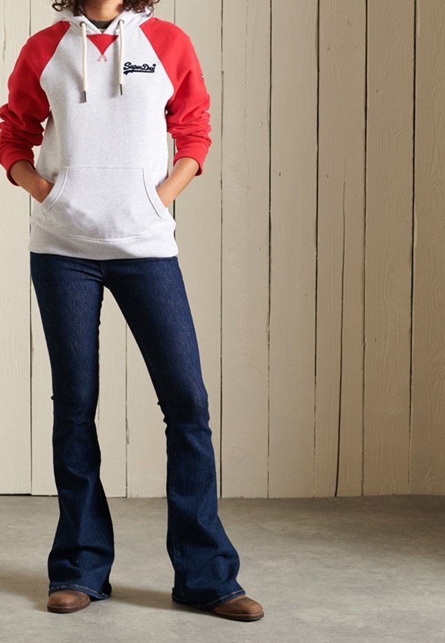 Personne portant un sweat à capuche blanc et rouge, un jean évasé foncé et des chaussures marron, debout contre un mur lambrissé en bois clair.