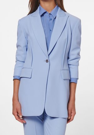 Vrouw draagt een lichtblauwe blazer over een bijpassend gebloemd shirt en broek, staand met ontspannen armen langs het lichaam.