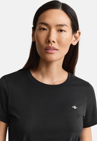 Γυναίκα με ίσια σκούρα μαλλιά που φοράει μαύρο μπλουζάκι με στρογγυλή λαιμόκοψη και μικρό λογότυπο GANT στο αριστερό στήθος, κοιτάζοντας μπροστά.