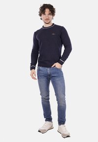 Maglione di filo blu navy con scollatura rotonda e polsini a costine, con dettagli bianchi e un piccolo logo sul petto, abbinato a jeans chiari e sneakers.