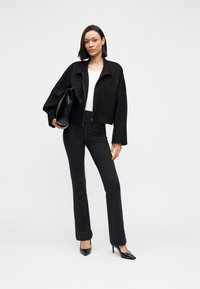 Jachetă neagră scurtă cu buzunare mari, top alb și pantaloni negri mulați. Modelul poartă o geantă neagră lucioasă și tocuri ascuțite.