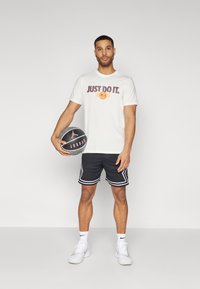 Hvit t-skjorte med "JUST DO IT" trykk og basketballgrafikk, sammen med svarte shorts med hvite detaljer, holder en basketball.