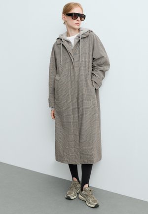 Personne portant un long manteau gris à carreaux avec un sweat à capuche en dessous, des leggings noirs, des baskets et de grandes lunettes de soleil foncées, debout contre un mur uni.