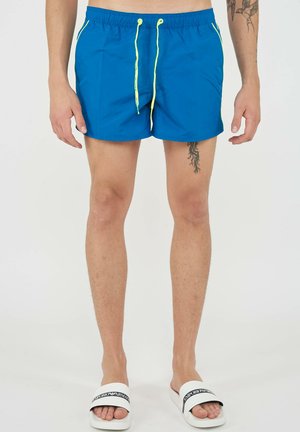 Man draagt blauwe zwemshorts met gele koordjes, witte slip-on sandalen en zichtbare tatoeages op het rechterbeen en de linkerarm.