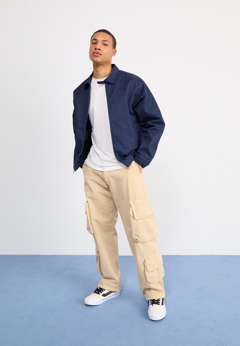 Veste bomber légère marine, t-shirt blanc, pantalon cargo beige avec poches et baskets blanches avec accents noirs. Tenue simple et décontractée.