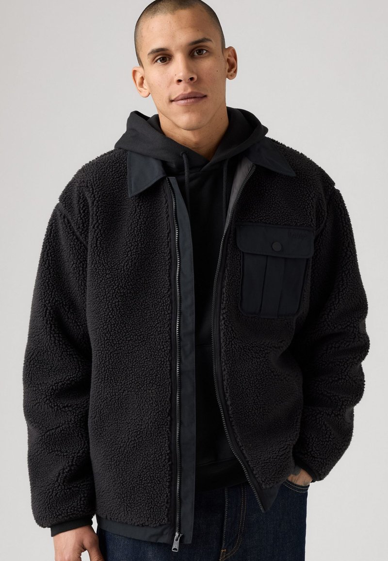 Sort fleece jakke med en glat krave, lynlås foran og en brystlomme. Laget over en sort hoodie med en struktureret, blød overflade.